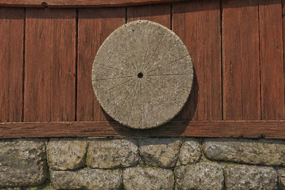 A round stone sun clock with carved hour lines, fixed to a red barn wall above a stone base.